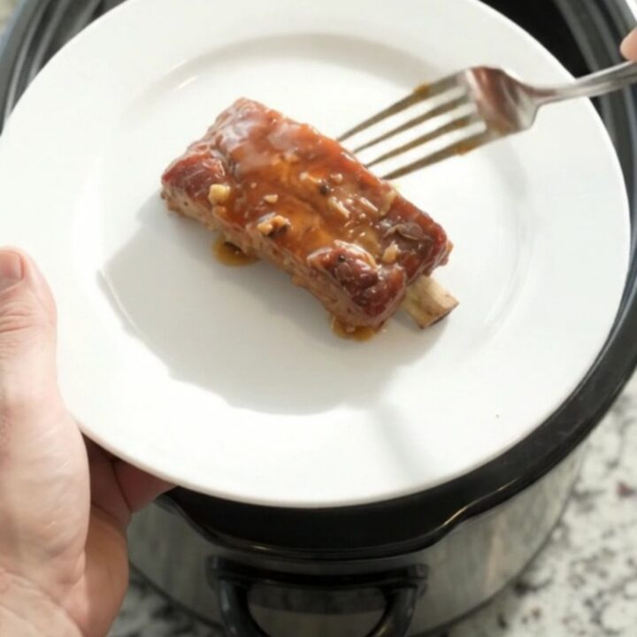Slow cooker filled with glazed pork ribs in maple bourbon sauce, topped with garlic and rosemary.