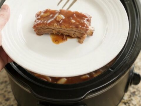 Root beer BBQ country ribs in a slow cooker, glazed with dark sauce and topped with caramelized onions.