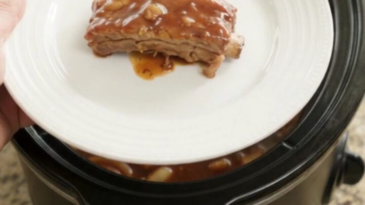 Root beer BBQ country ribs in a slow cooker, glazed with dark sauce and topped with caramelized onions.