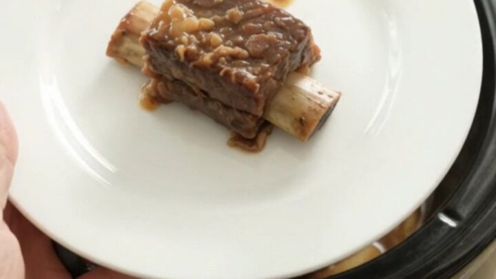 A glossy, caramelized beef short rib on a white plate, with the slow cooker in the background