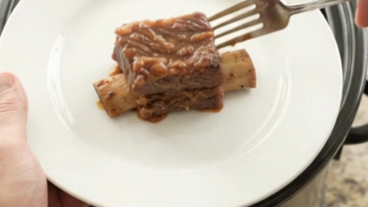Glossy BBQ short ribs in a black slow cooker, garnished with rosemary