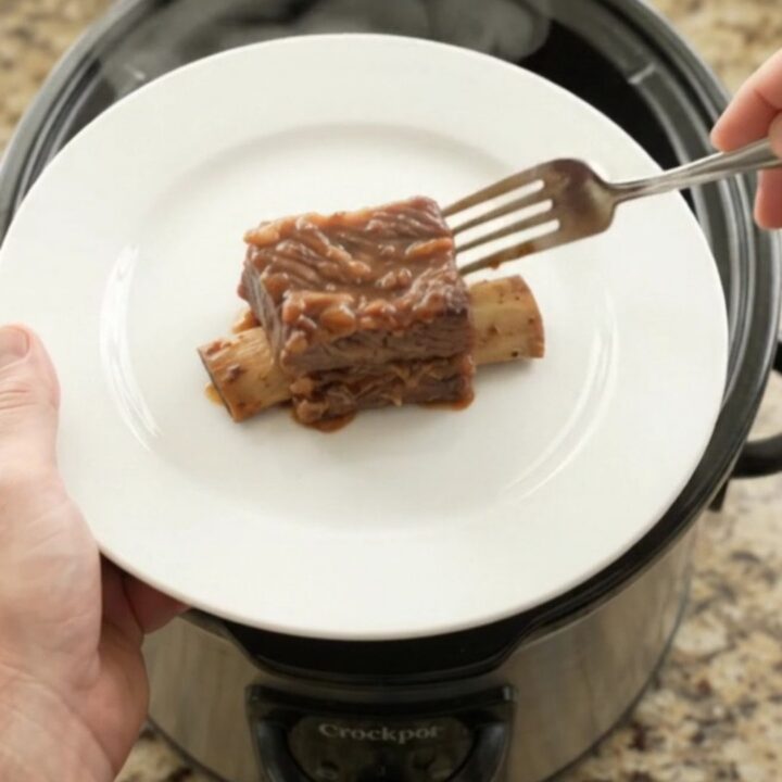Glossy BBQ short ribs in a black slow cooker, garnished with rosemary