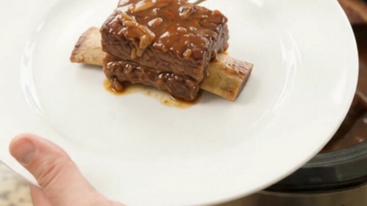 A slow cooker filled with glistening BBQ short ribs in a rich dark sauce, steam rising, on a kitchen counter.