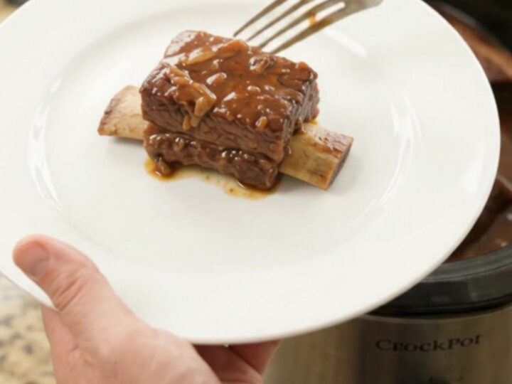 A slow cooker filled with glistening BBQ short ribs in a rich dark sauce, steam rising, on a kitchen counter.