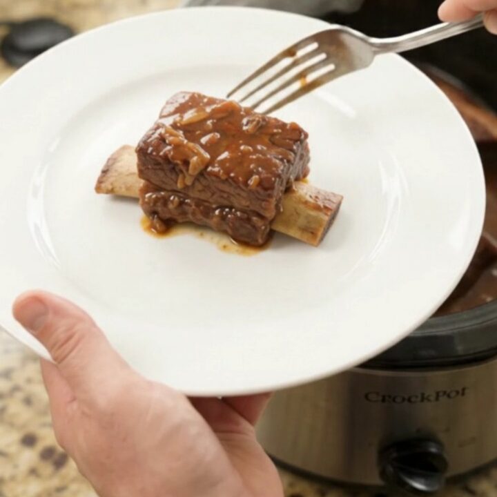 A slow cooker filled with glistening BBQ short ribs in a rich dark sauce, steam rising, on a kitchen counter.