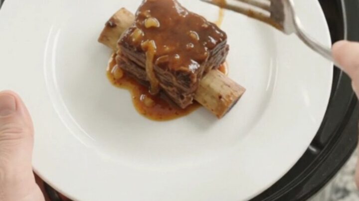 A pile of glistening BBQ short ribs on a white plate, garnished with caramelized onions
