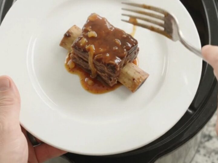 A pile of glistening BBQ short ribs on a white plate, garnished with caramelized onions