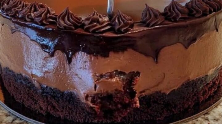 A decadent chocolate mousse cake with a glossy ganache glaze and chocolate rosettes on a granite countertop.