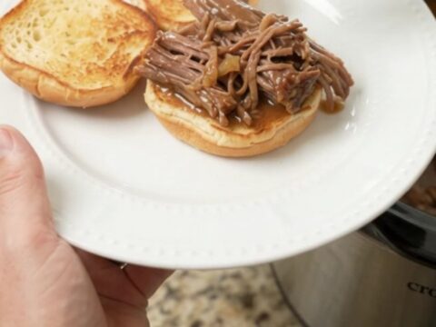 Shredded beef chuck roast in a glossy brown gravy inside a black slow cooker insert, steam rising.