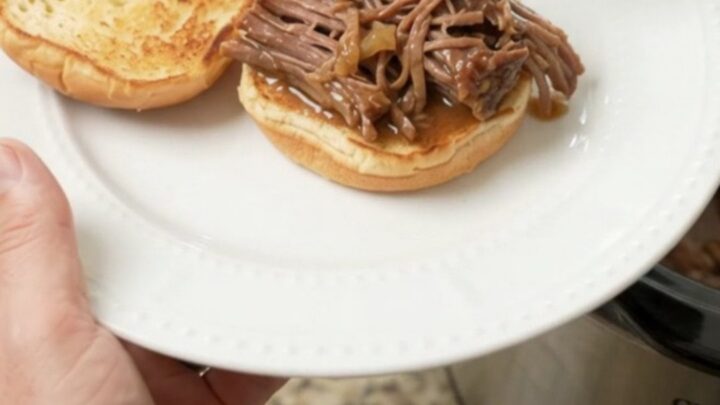 Shredded beef chuck roast in a glossy brown gravy inside a black slow cooker insert, steam rising.