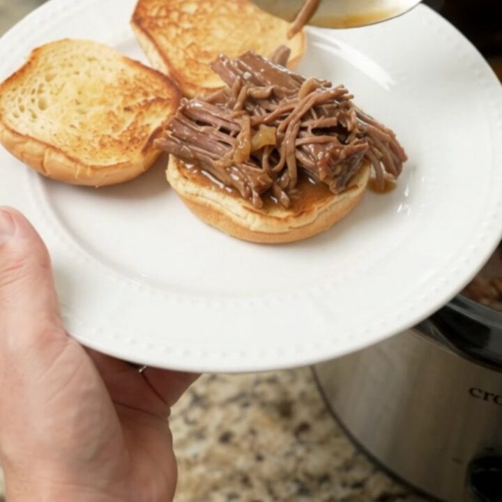 Shredded beef chuck roast in a glossy brown gravy inside a black slow cooker insert, steam rising.