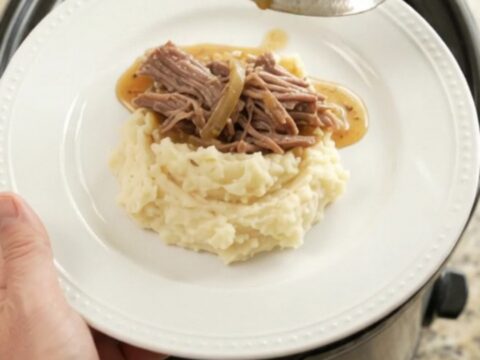 Shredded beef chuck roast with caramelized onions and buttery jus in a slow cooker, topped with a spoonful of sauce.