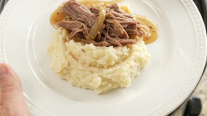 Shredded beef chuck roast with caramelized onions and buttery jus in a slow cooker, topped with a spoonful of sauce.