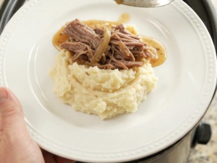 Shredded beef chuck roast with caramelized onions and buttery jus in a slow cooker, topped with a spoonful of sauce.