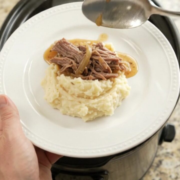 Shredded beef chuck roast with caramelized onions and buttery jus in a slow cooker, topped with a spoonful of sauce.