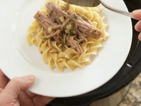 A slow cooker filled with shredded pork shoulder in a rich garlic-herb gravy, with a spoon lifting a steaming mound.