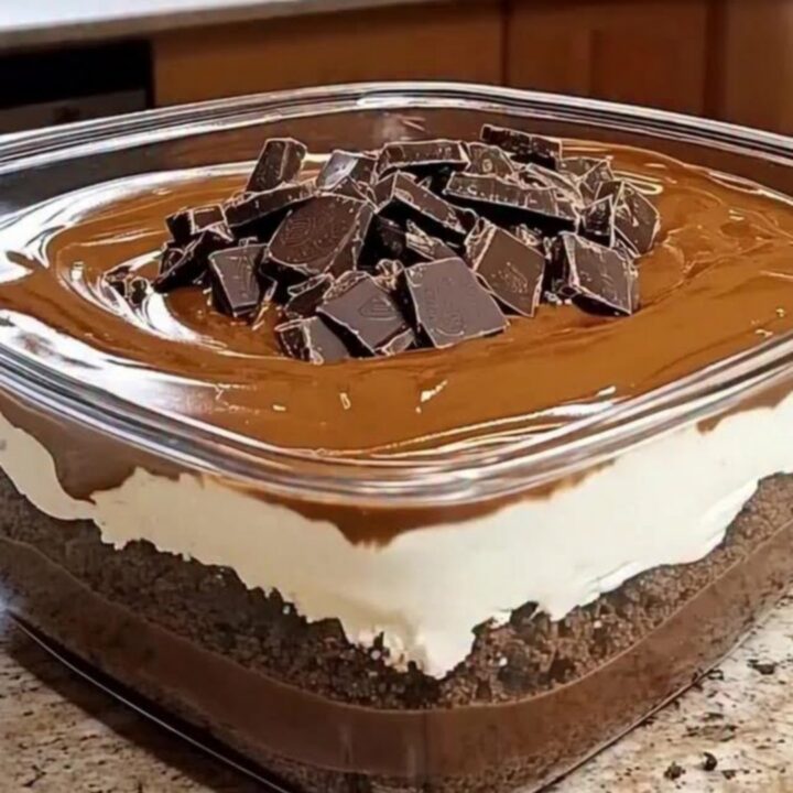 A close-up of a square glass dish filled with layers of chocolate pudding, cookie crumbs, cream, and ganache.