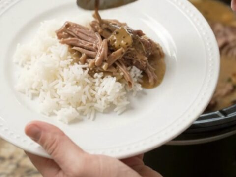 Shredded pork sirloin roast in creamy Dijon gravy in a black slow cooker, garnished with fresh herbs.