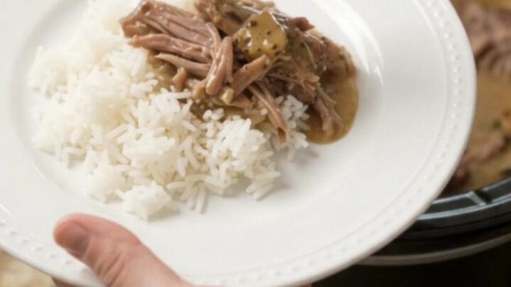 Shredded pork sirloin roast in creamy Dijon gravy in a black slow cooker, garnished with fresh herbs.