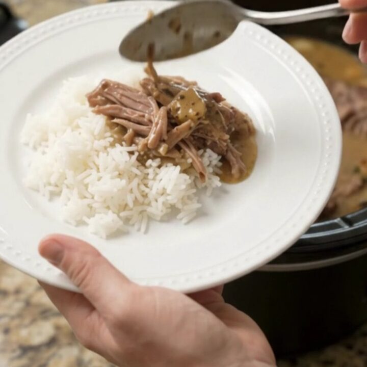 Shredded pork sirloin roast in creamy Dijon gravy in a black slow cooker, garnished with fresh herbs.