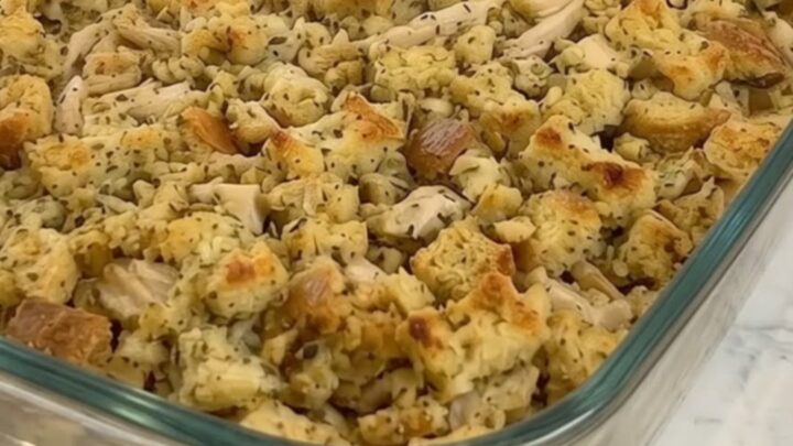 A glass baking dish filled with golden-brown chicken and herb stuffing casserole on a white marble countertop.