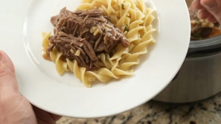 Slow cooker beef roast with garlic and herbs in a black Crockpot, topped with butter medallions.