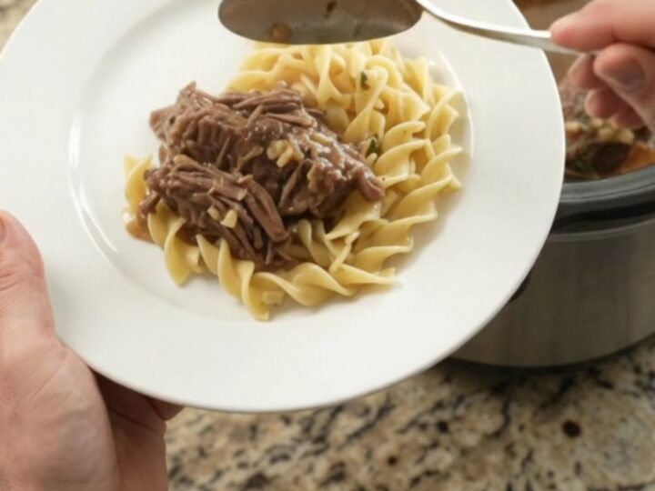 Slow cooker beef roast with garlic and herbs in a black Crockpot, topped with butter medallions.