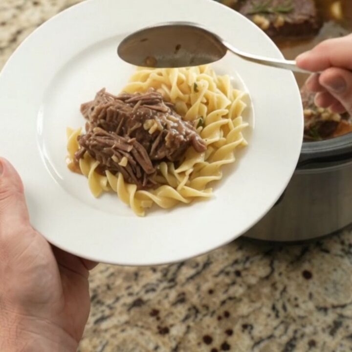 Slow cooker beef roast with garlic and herbs in a black Crockpot, topped with butter medallions.