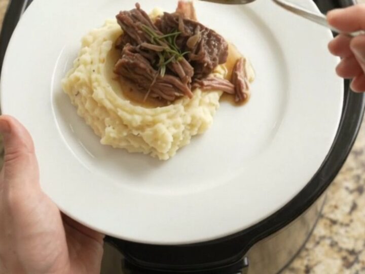 A fork-tender bottom round roast in a slow cooker with garlic and herbs, golden jus bubbling around it.