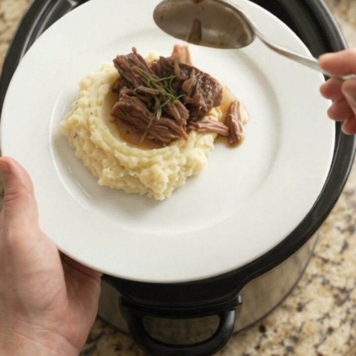 A fork-tender bottom round roast in a slow cooker with garlic and herbs, golden jus bubbling around it.