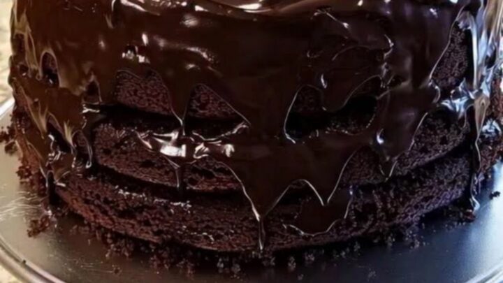 A towering three-layer chocolate fudge cake on a silver pedestal stand with glossy ganache.