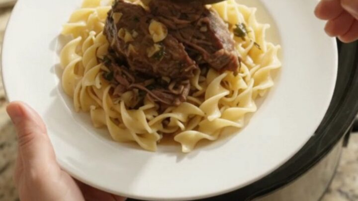 A slow cooker filled with shredded beef roast, shallots, and carrots in a buttery sauce, with steam rising.
