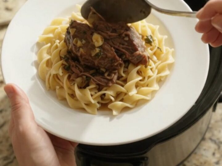 A slow cooker filled with shredded beef roast, shallots, and carrots in a buttery sauce, with steam rising.