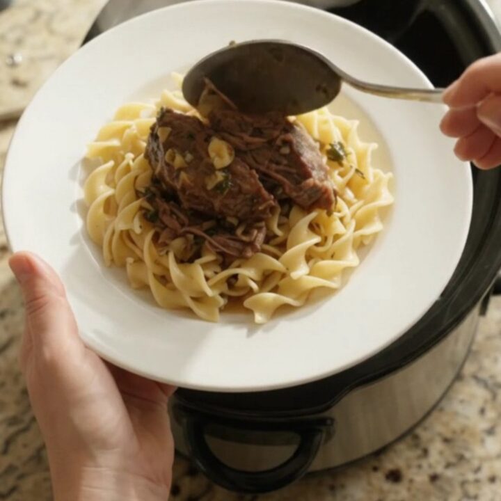 A slow cooker filled with shredded beef roast, shallots, and carrots in a buttery sauce, with steam rising.