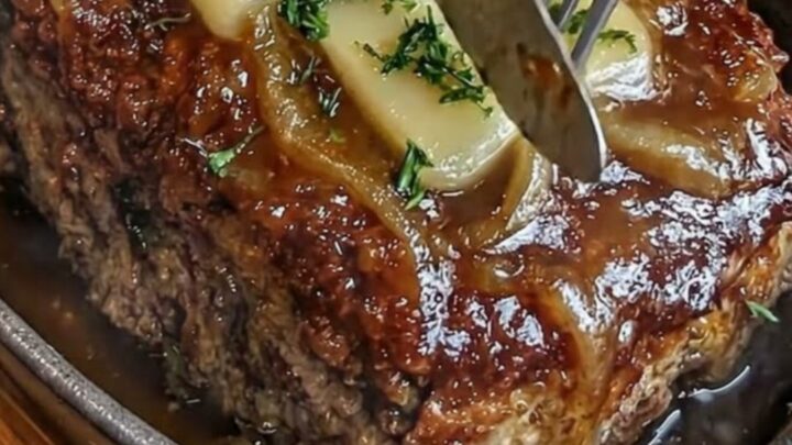A close-up of a French onion meatloaf in a cast-iron skillet, topped with melted Swiss cheese and caramelized onions.