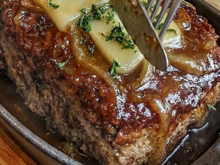 A close-up of a French onion meatloaf in a cast-iron skillet, topped with melted Swiss cheese and caramelized onions.