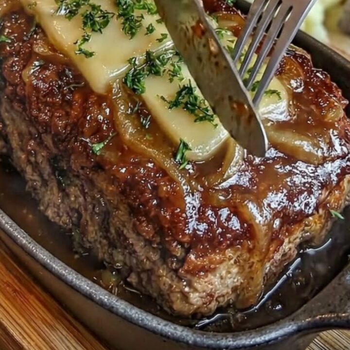 A close-up of a French onion meatloaf in a cast-iron skillet, topped with melted Swiss cheese and caramelized onions.
