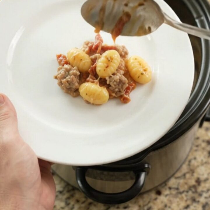 Crockpot Parmesan Sausage Tuscan Gnocchi in a black slow cooker, topped with melted cheese and steam rising.
