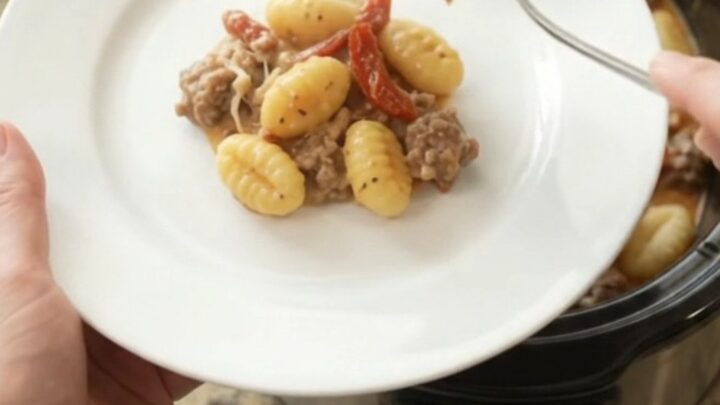 A slow cooker filled with creamy sausage gnocchi, topped with melted provolone and sun-dried tomatoes.