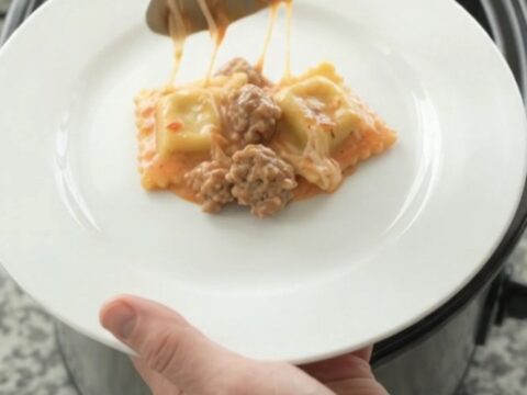 A slow cooker filled with cheesy sausage vodka ravioli, with melted mozzarella and pink sauce bubbling.