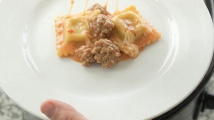 A slow cooker filled with cheesy sausage vodka ravioli, with melted mozzarella and pink sauce bubbling.