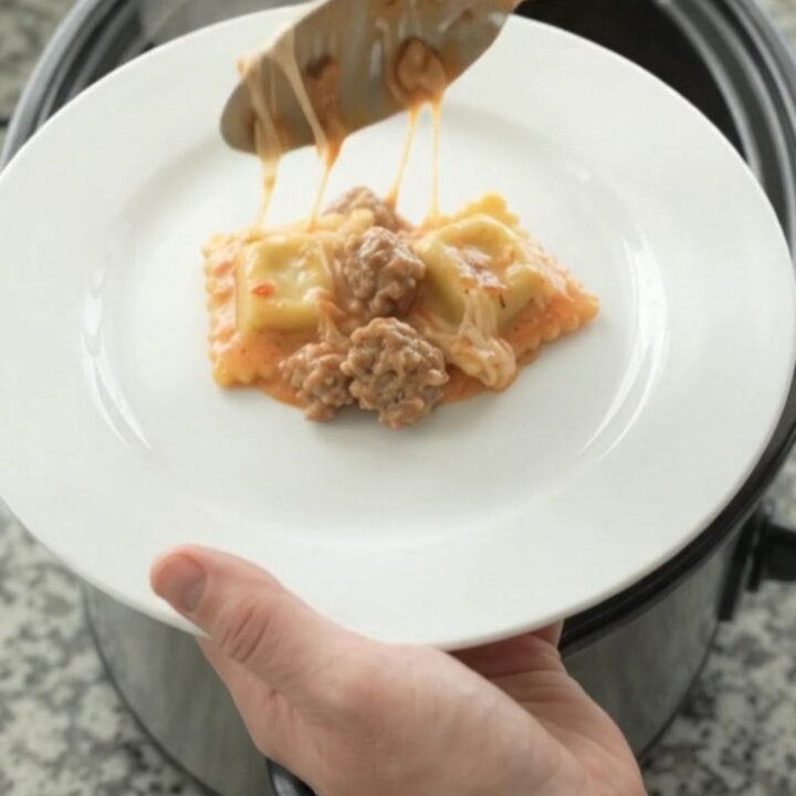 A slow cooker filled with cheesy sausage vodka ravioli, with melted mozzarella and pink sauce bubbling.