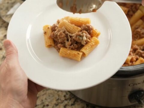 Slow cooker filled with creamy rigatoni, sausage, and melted cheese, topped with herbs.