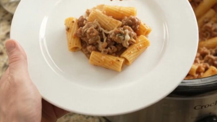 Slow cooker filled with creamy rigatoni, sausage, and melted cheese, topped with herbs.