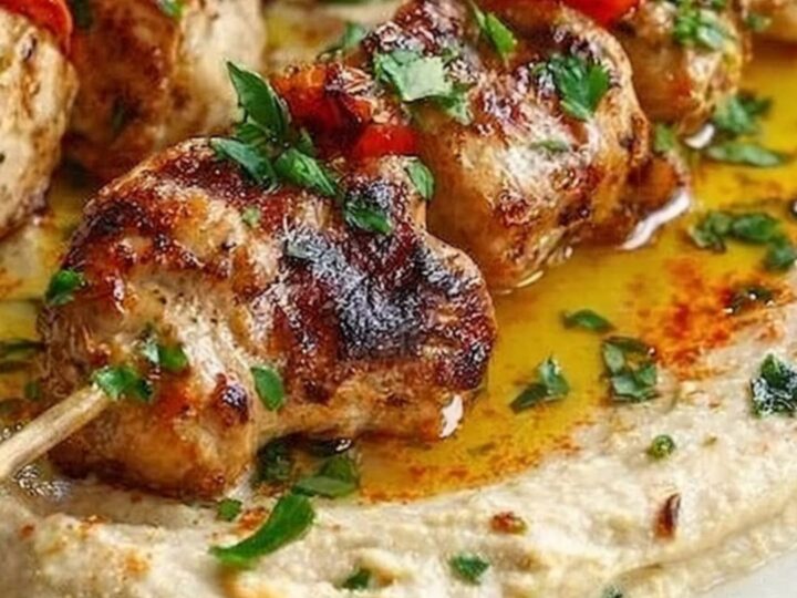 A rustic wooden table featuring grilled chicken skewers served over a smooth bed of hummus.