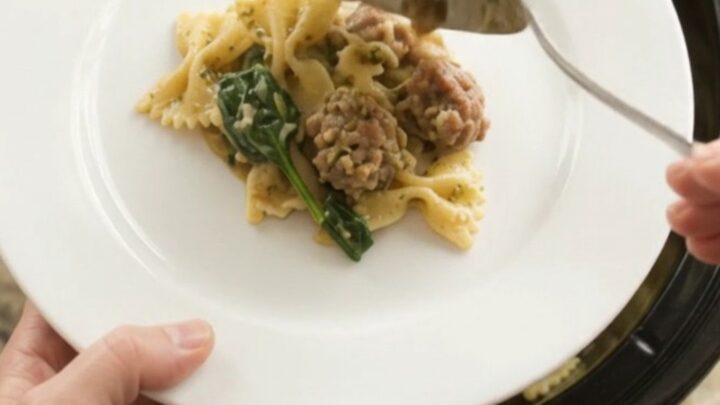 Slow cooker Parmesan sausage pesto farfalle in a black crockpot with melted cheese.