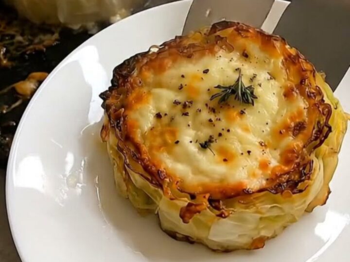 A close-up of a golden-brown cabbage steak topped with melted, bubbling cheese and fresh thyme on a white plate.