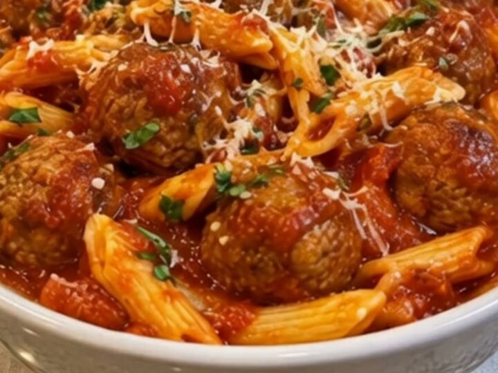 A white ceramic bowl filled with penne pasta and juicy meatballs in marinara sauce topped with parmesan and parsley.
