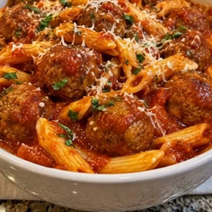 A white ceramic bowl filled with penne pasta and juicy meatballs in marinara sauce topped with parmesan and parsley.