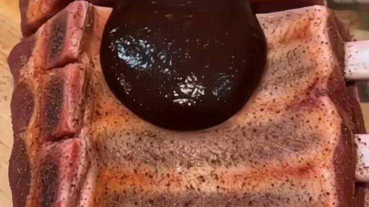 A close-up shot of glossy, dark barbecue sauce being ladled over tender, slow-cooked ribs on a wooden cutting board.